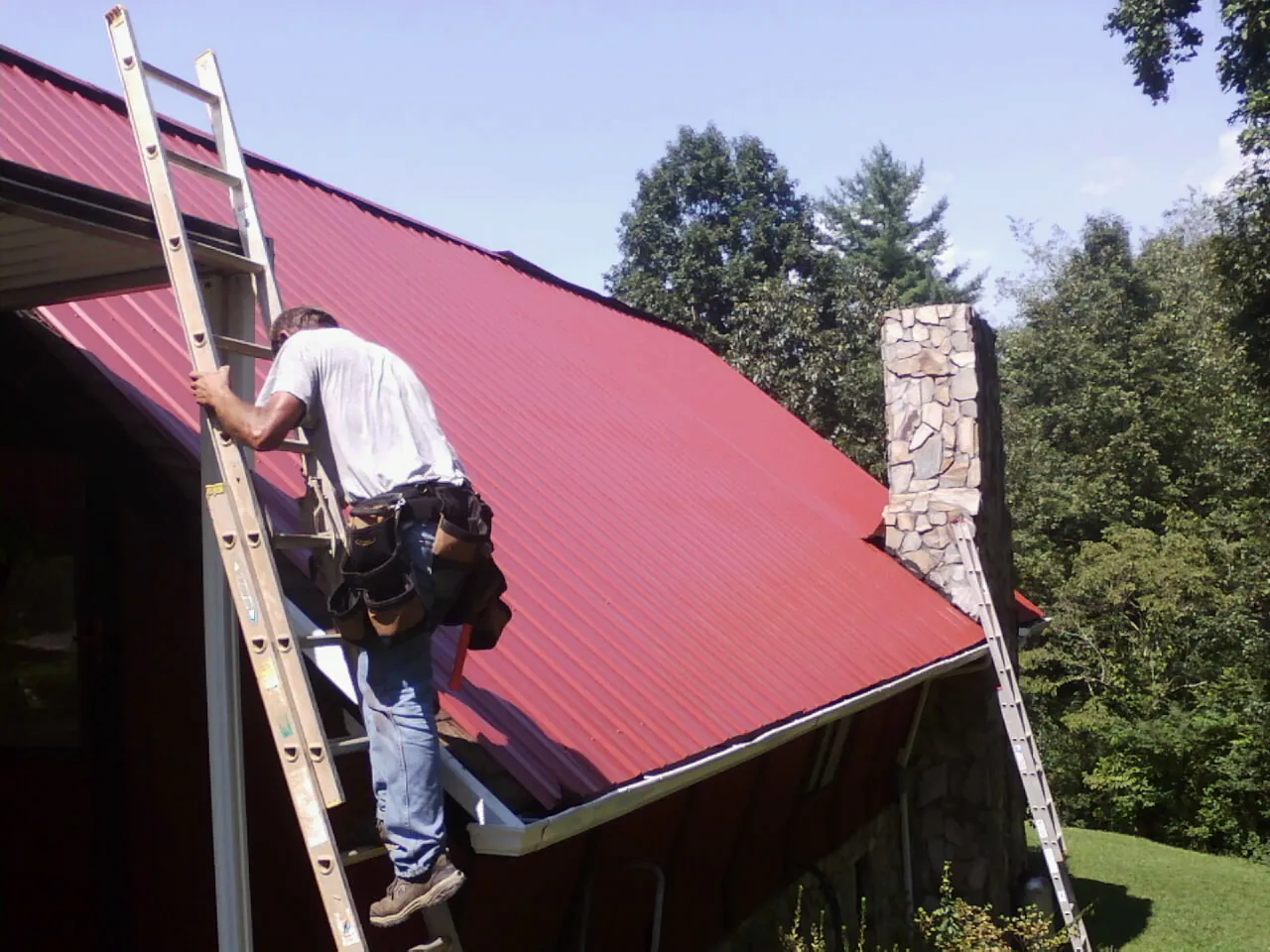 Roofer on a ladder beside a red metal roof and stone chimney — Soffit & Fascia Repair by Roofing Brothers in Aurora